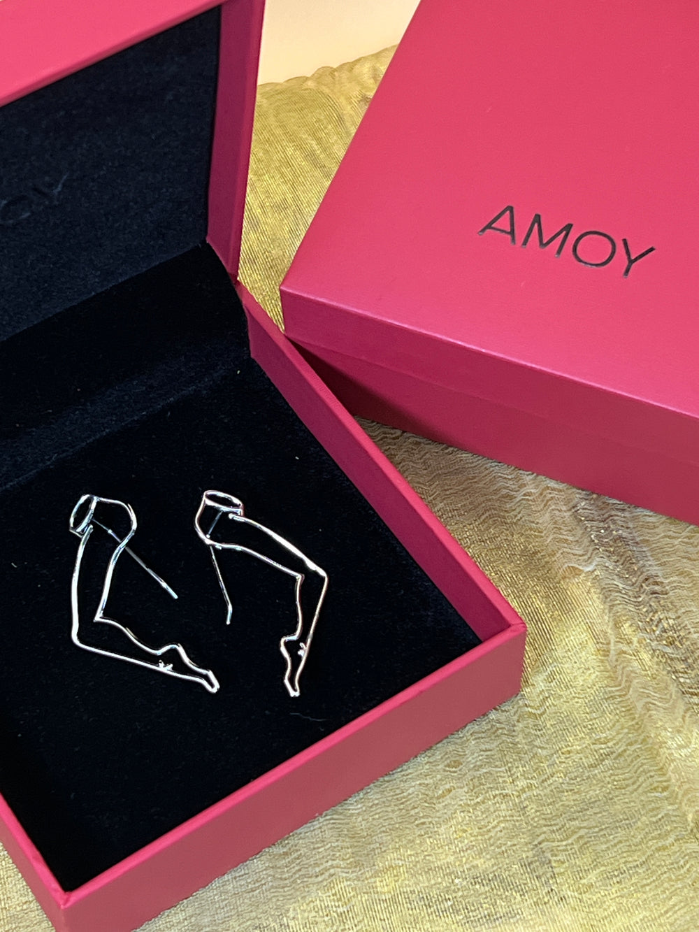 Silver wire earrings shaped like minimalist sketch-style legs in a pink AMOY box with black interior lining.